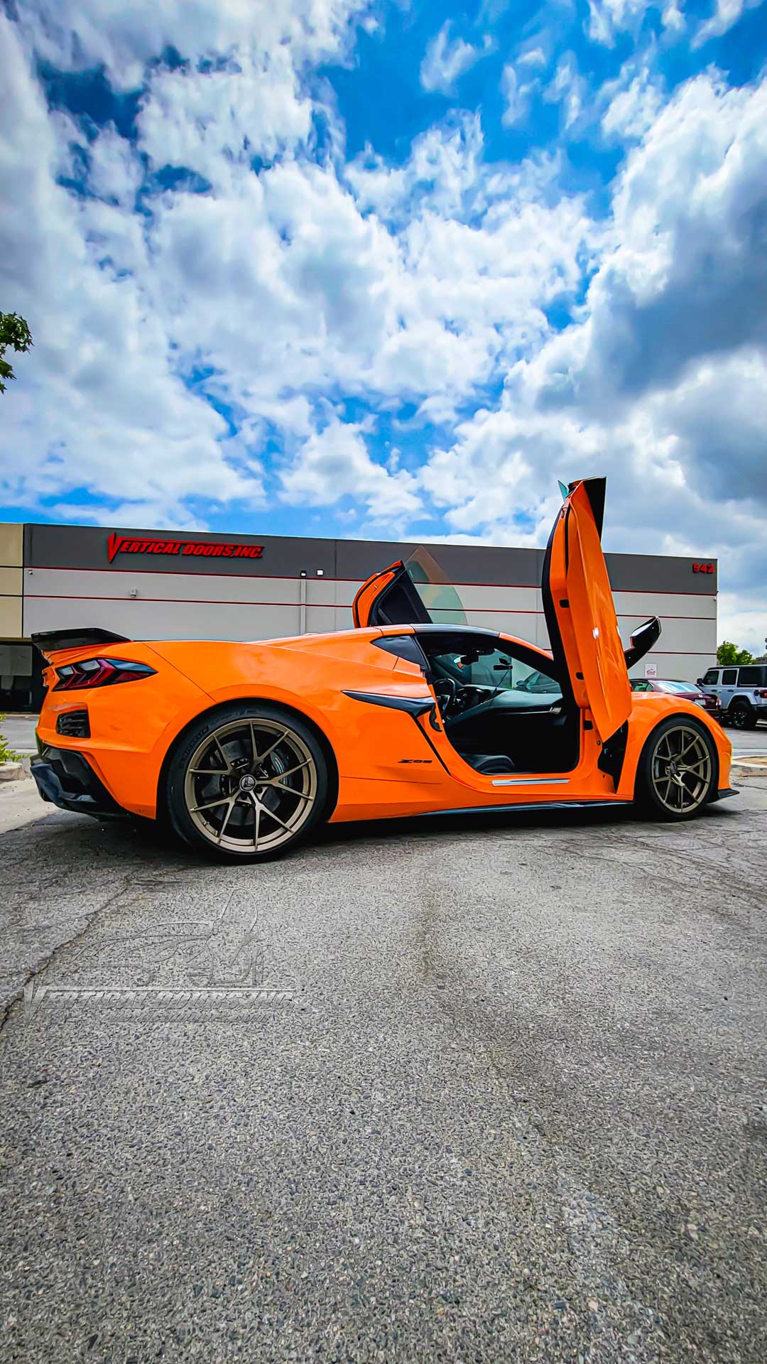 Orange sports car with C8 Z06 vertical door kit installed and doors open in front of Vertical Doors Inc building
