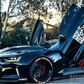 Black Chevrolet Camaro with vertical doors installed using Chevrolet Camaro vertical door kit in a wooded driveway